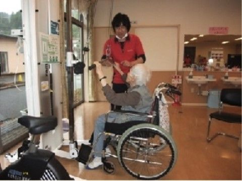 車いすに座りながら上半身用のトレーニングマシンを使用する入居者と指導するスタッフ