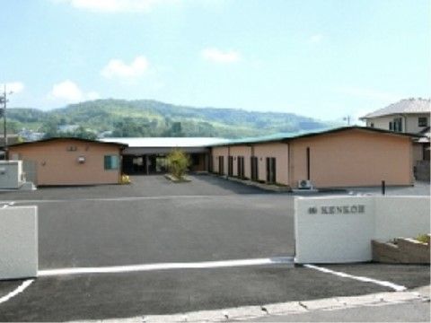 施設の門から見える広い敷地とベージュ色のコの字型の平屋建ての建物