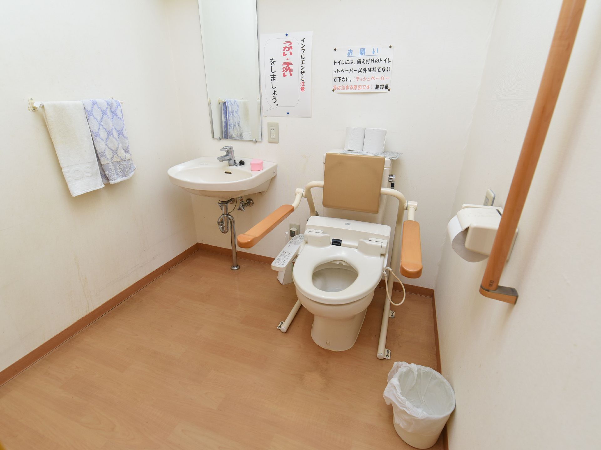 フローリング調の床に白い壁の清潔感のあるトイレである。手洗いスペースと手すり、呼び出しボタンが設置されている。