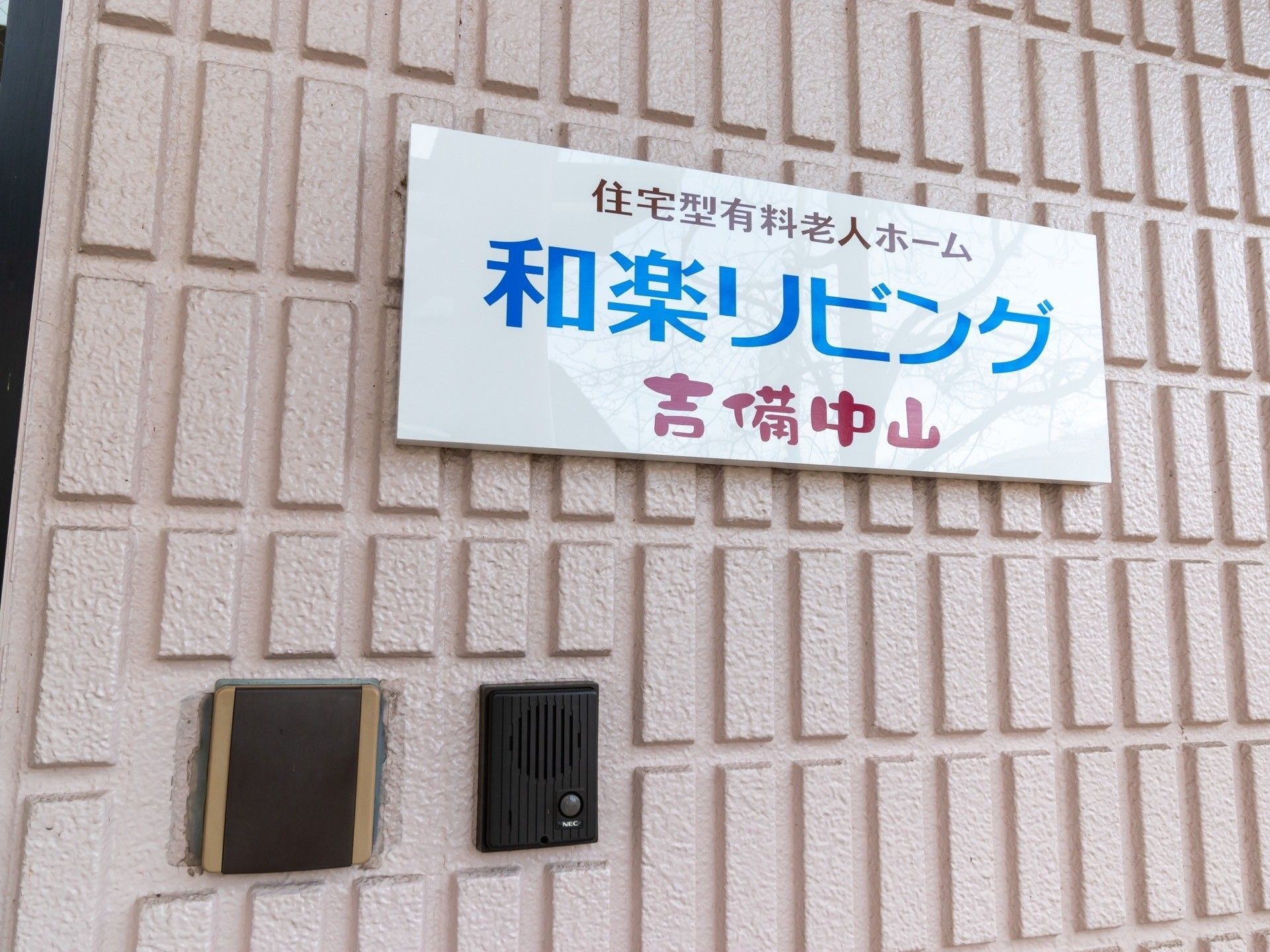 白地に青い字で施設名が書かれた看板が掲げられている。壁面はブロック状になっており、インターホンが設けられている。