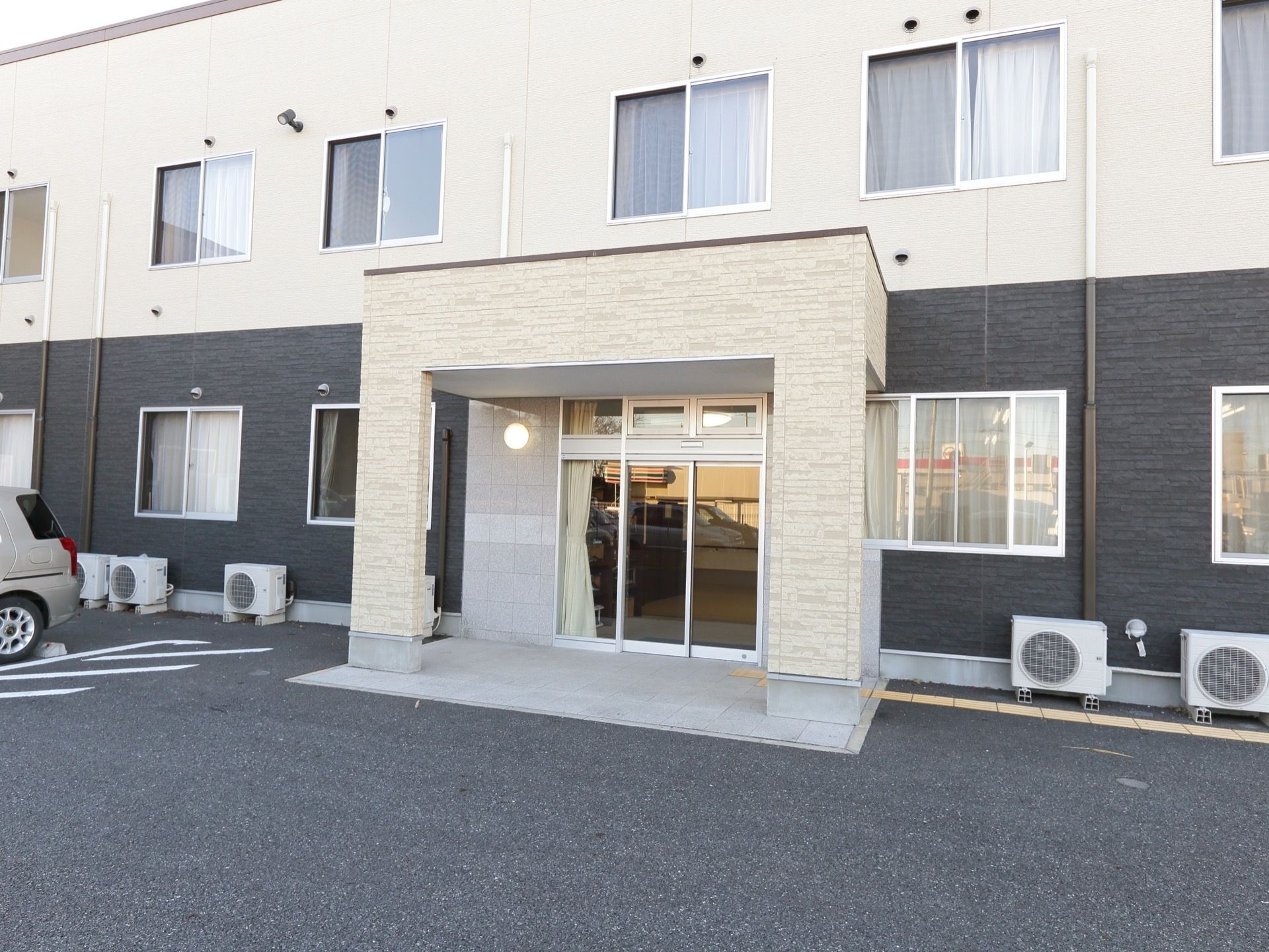 ダークグレーとベージュの外壁が落ち着いた雰囲気になっている施設の外観。入り口には屋根が設置されている。