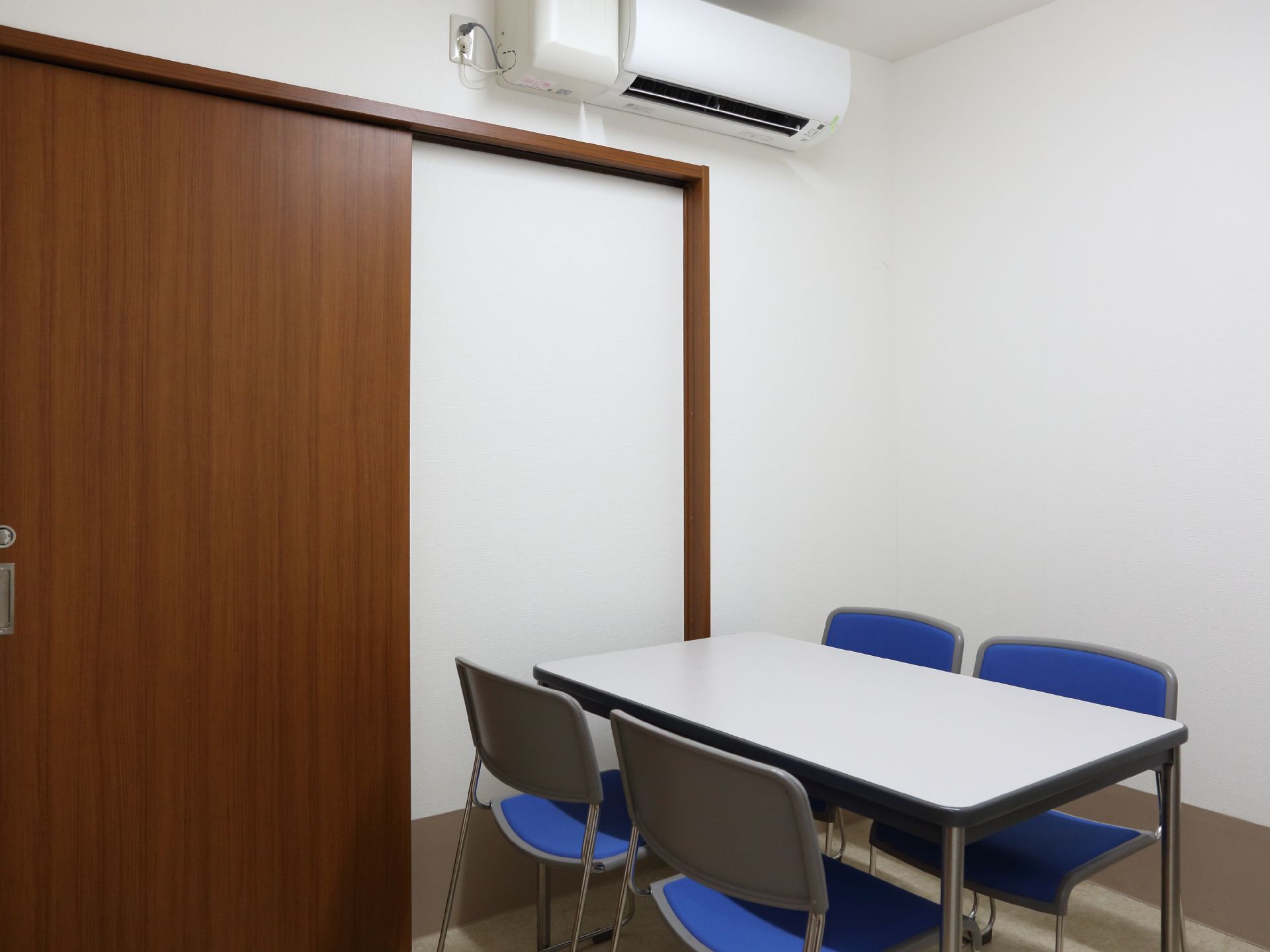 面会室の中には、白いテーブルが1台と青色の椅子が4脚置かれている。室内には、エアコンが完備されている。