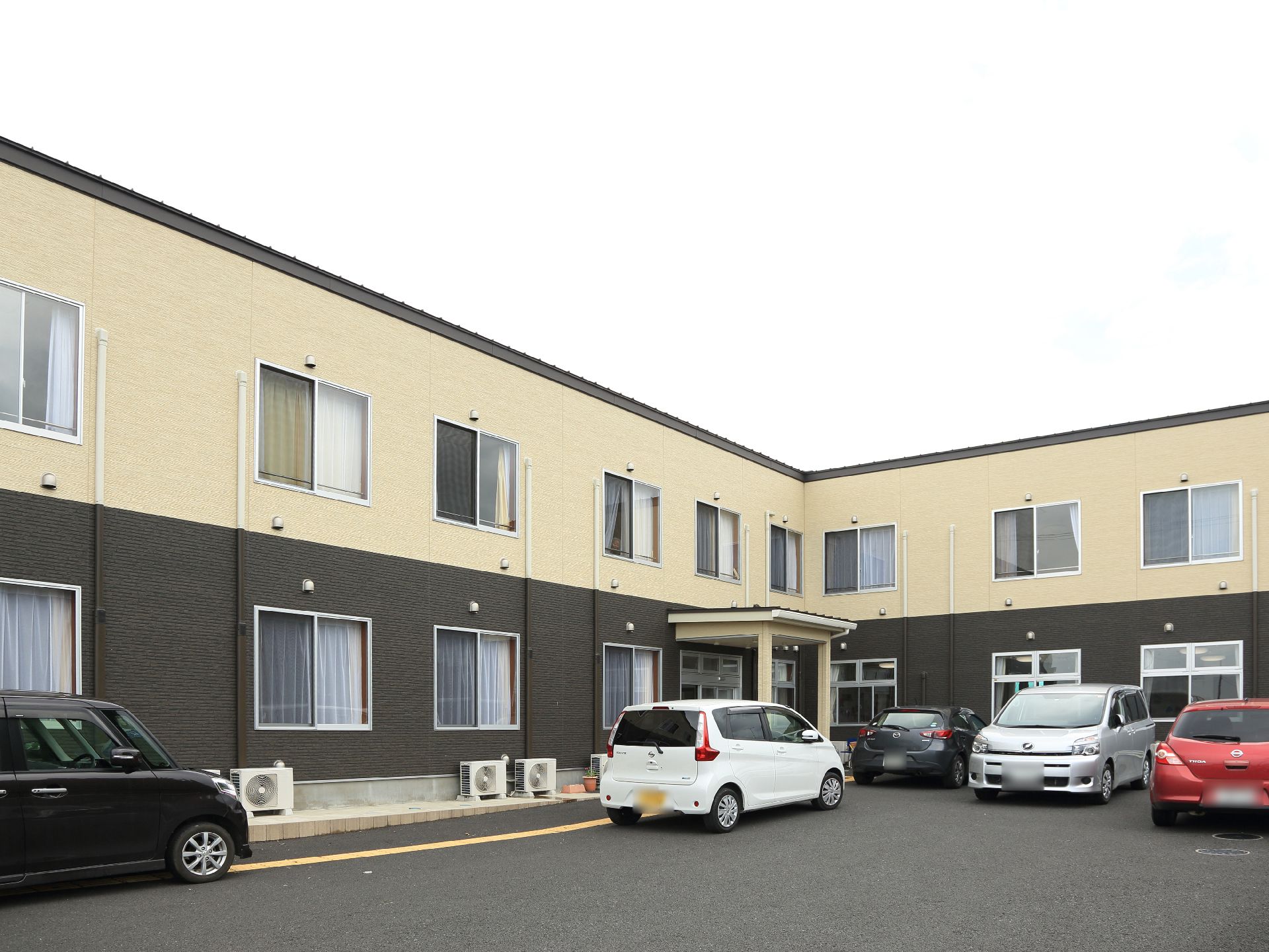 施設の建物は、2階建てである。外壁の色が、2階はベージュ、1階は黒色をしている。建物の前には、駐車スペースがある。