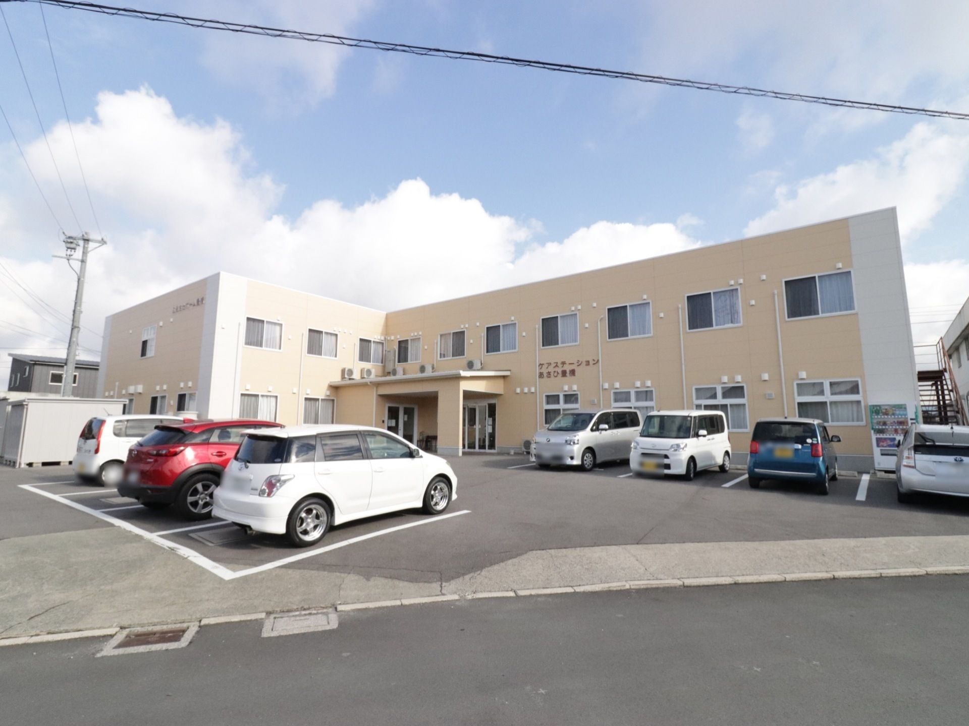 ベージュを基調とした２階建ての外観である。中央の外壁には、ダークブラウンの文字で施設名を表示している。敷地内には駐車スペースがある。