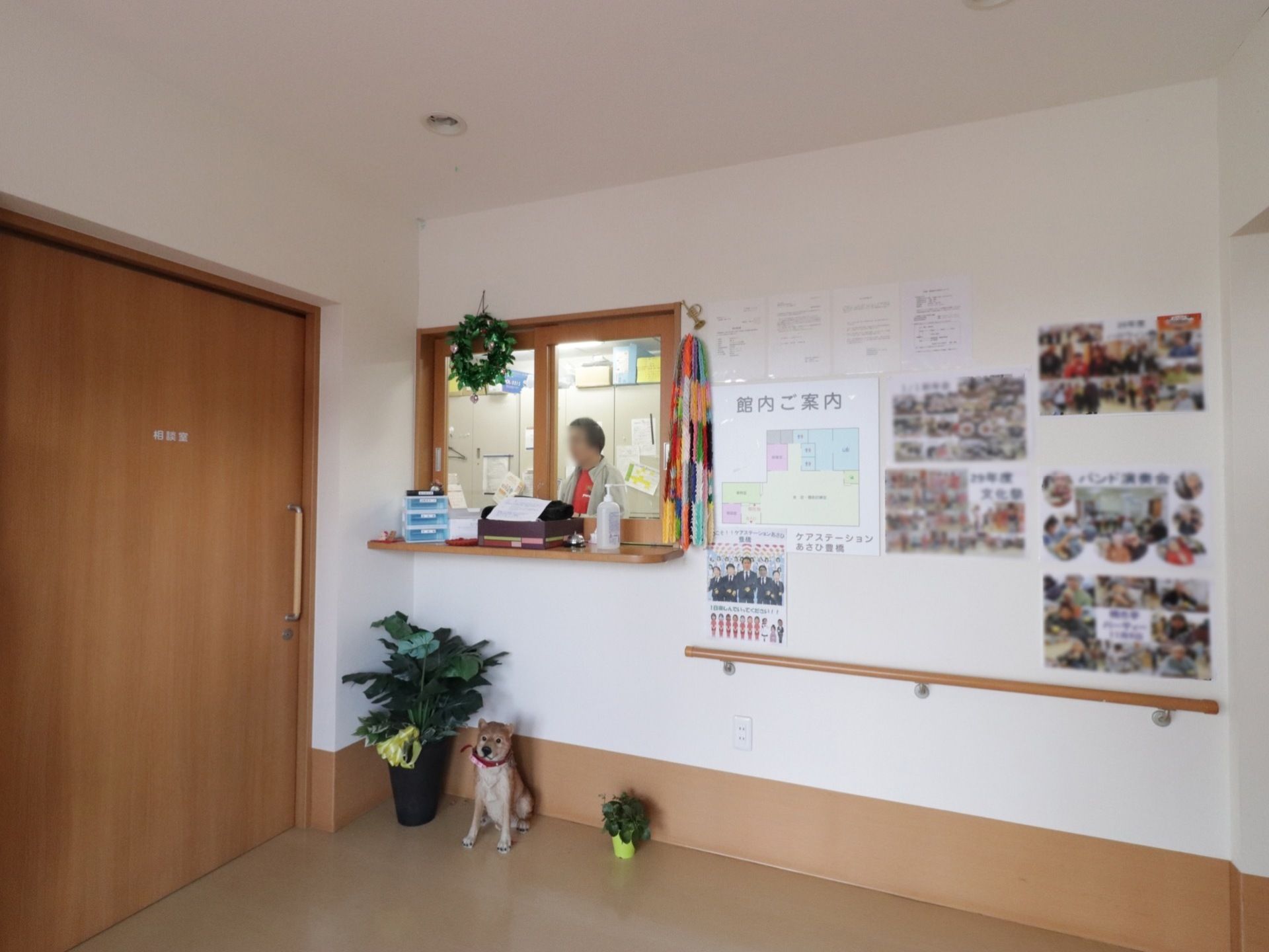 玄関出入口正面には、スタッフルームの窓口がある。下には観葉植物や犬の置物を置いている。壁には館内の案内図や写真を貼っている。