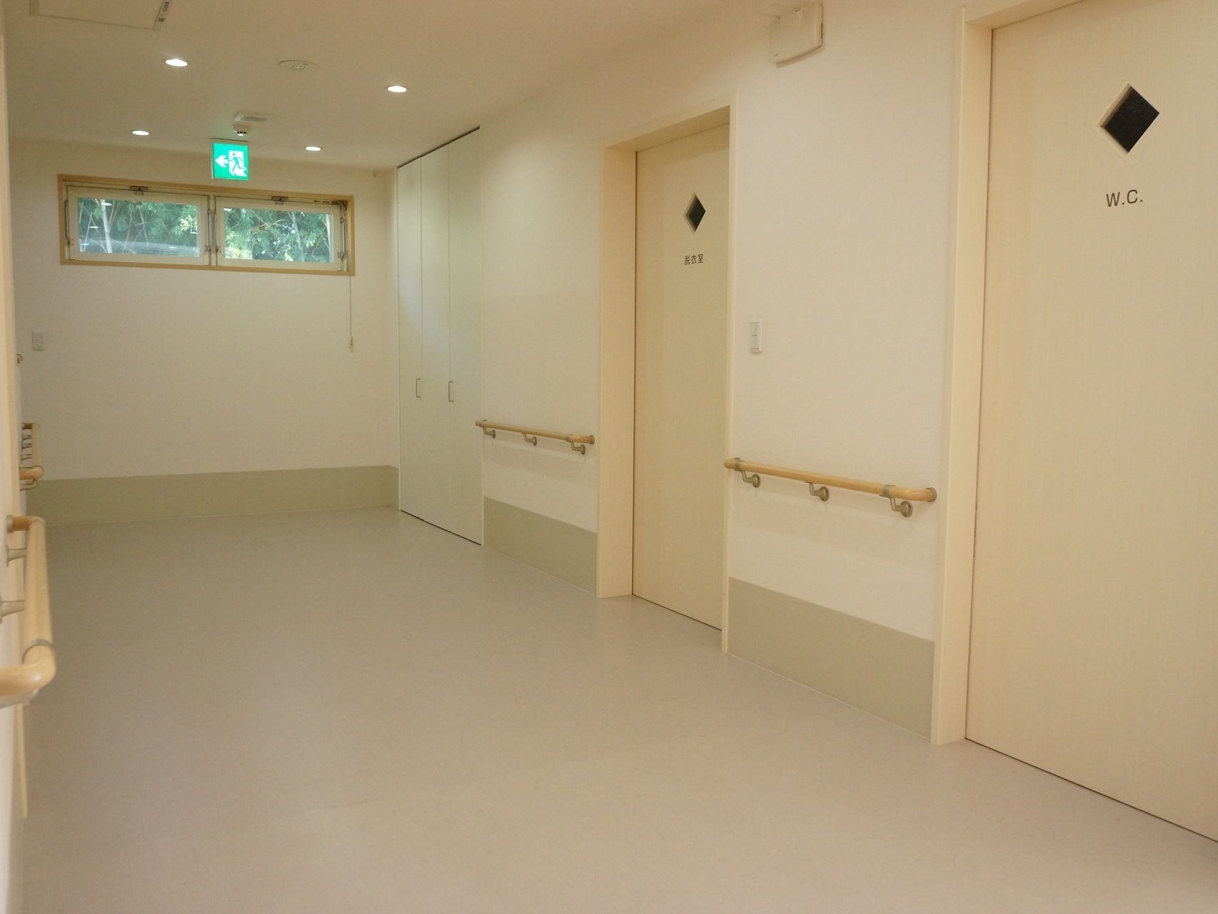 施設二階の廊下である。右の壁側に、収納、脱衣室とトイレの扉が並んでいる。壁には手すりが設けられている。