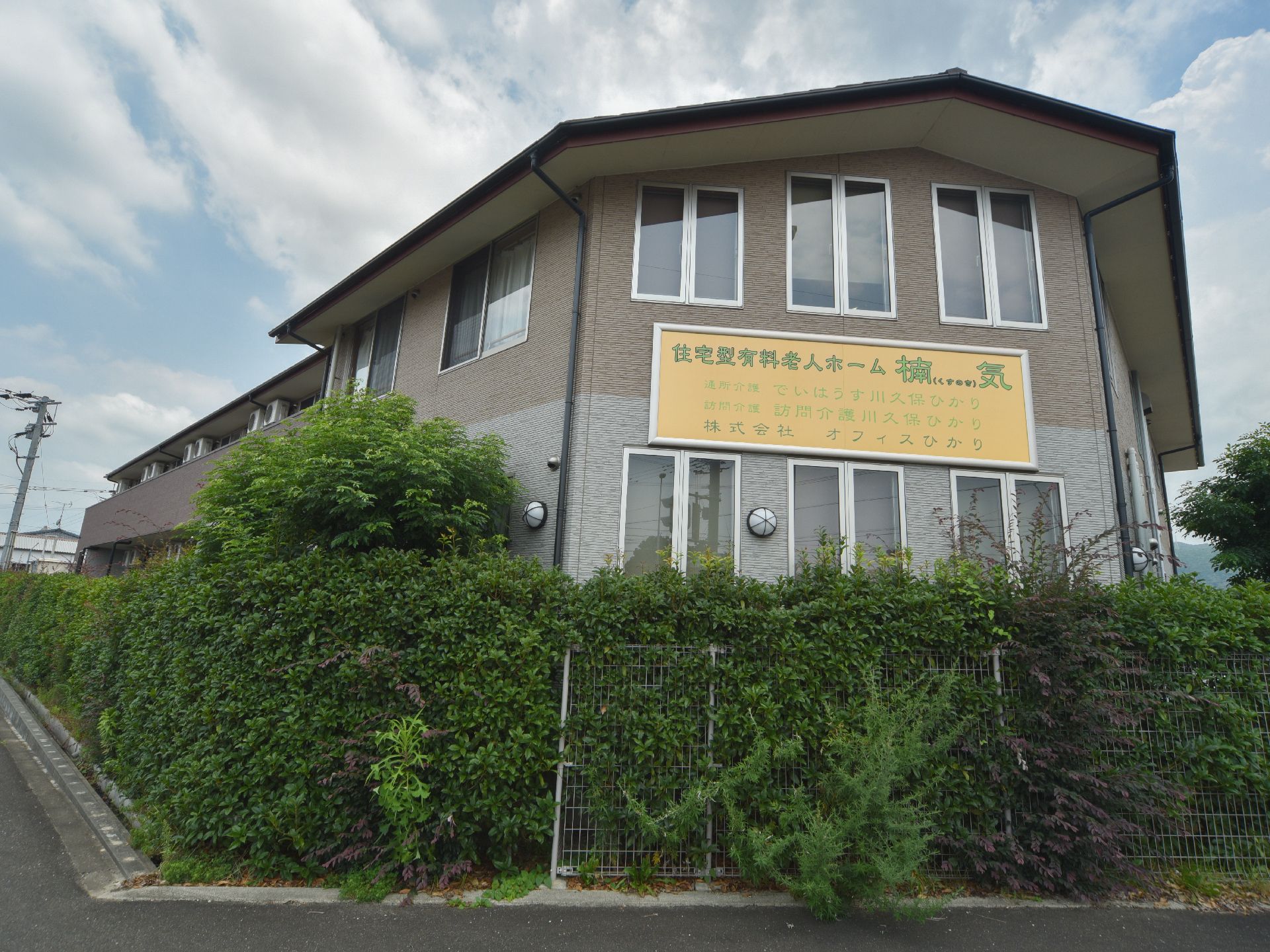 手前が緑に囲まれている施設外観が映し出されており、壁面には施設名の書かれた看板が黄色で目立つように掲げられている。