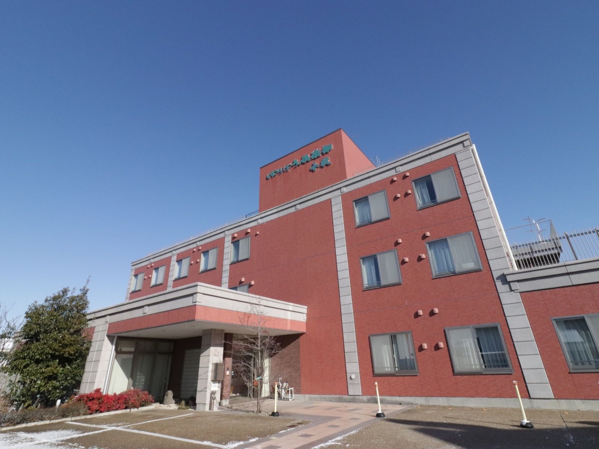 レンガ色の壁に緑の文字で「ゆうゆう倶楽部」と描かれた建物。玄関まで段差の無いフラットな造り。玄関の周りには、植物が植えられている。