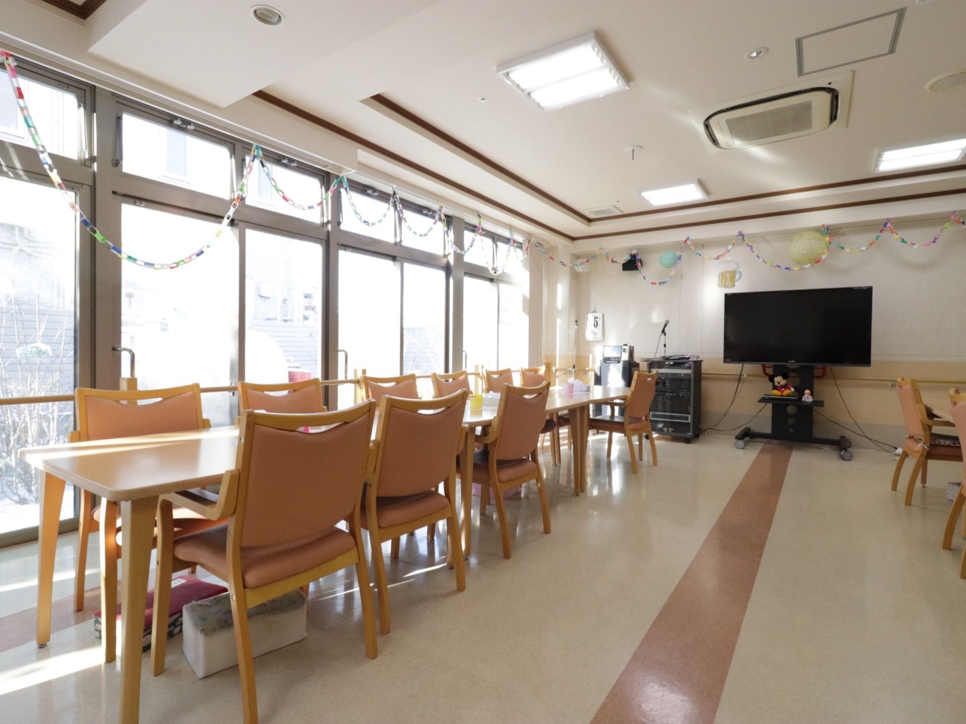 長い机に椅子が並んでいる。窓が大きく、明るい日差しが差し込んでいる。また、テレビとカラオケの機械が置かれている。
