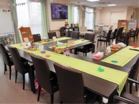 窓から光が入る、広くて明るい室内。手前には長机がコの字型に並ぶ。奥は壁掛けの大画面テレビに向かい、10脚ほどの椅子が並ぶ。