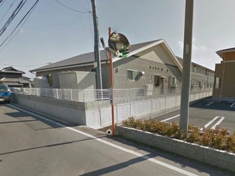 道路に面して低いフェンスに囲まれた新築の２階建。周囲には一般の住宅があるが、視界が開けており周囲にも溶け込んでいる。