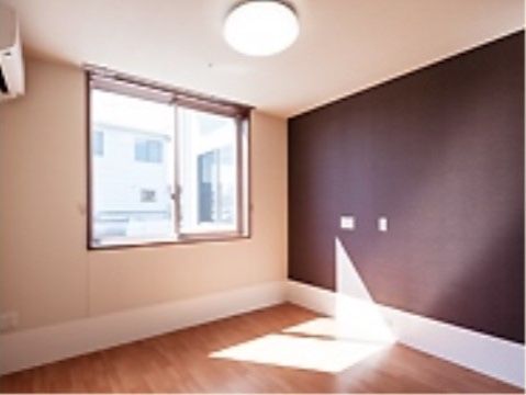腰高窓があり、照明は丸いシーリングライトである。エアコンが壁の上部にある。床は明るい木目調で窓の壁と天井はアイボリーである。