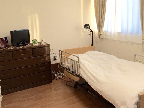 介護用の手すりがついたベッドの横に、小さなタンスがおいてある。タンスの上には、テレビや時計、小物などが並んでいる。