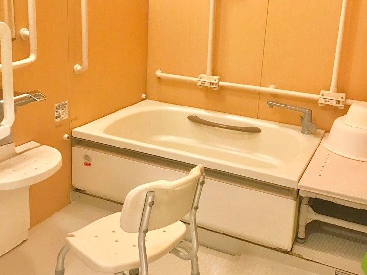 浴槽、シャワー、背もたれ付きのシャワーチェアがあり、黄色系の壁に白い手すりがたくさん設置された浴室