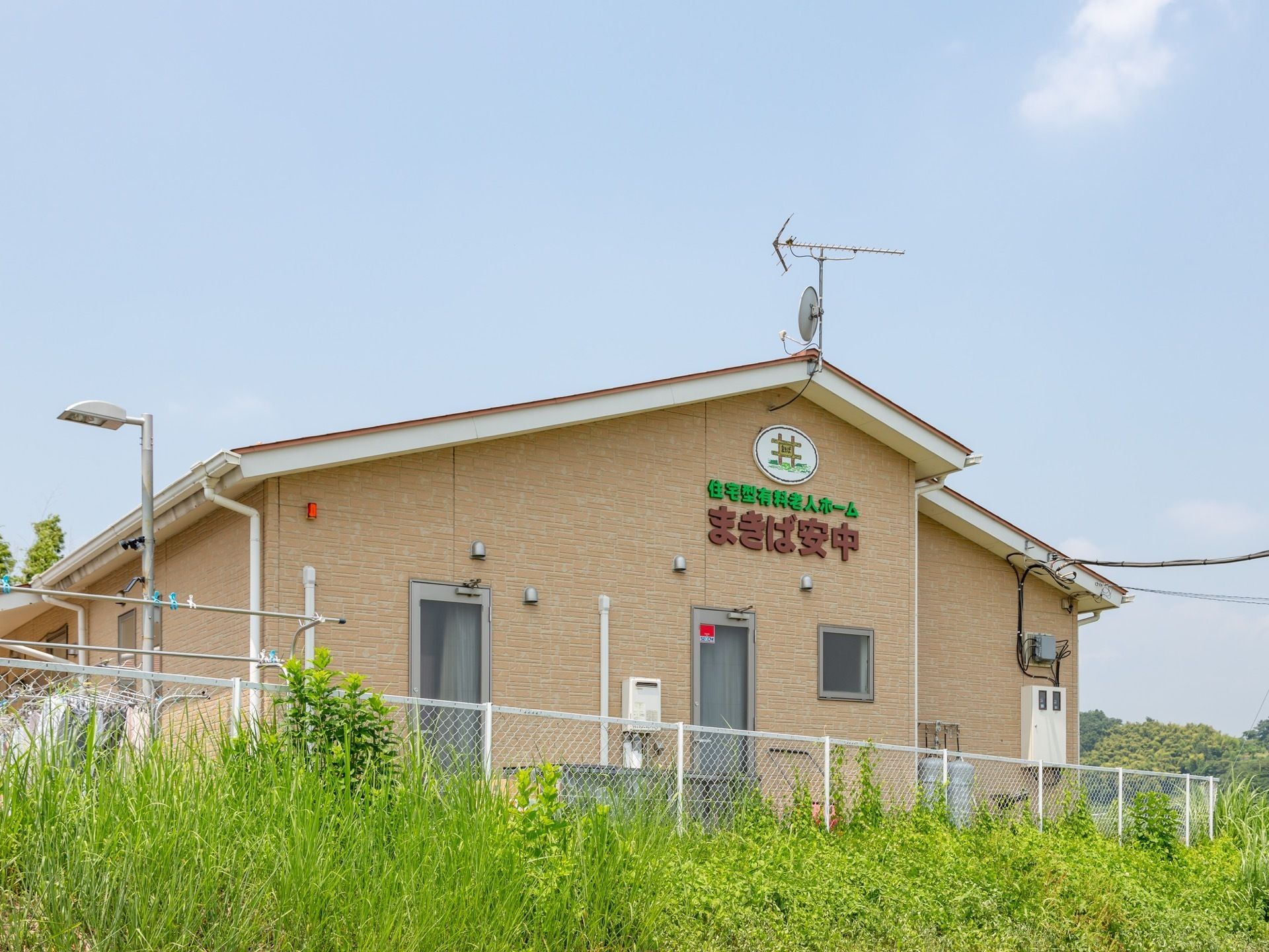 優しいベージュ色の平屋建てである。外壁には当施設名が掲げられている。敷地の周囲は白いフェンスで囲われている。