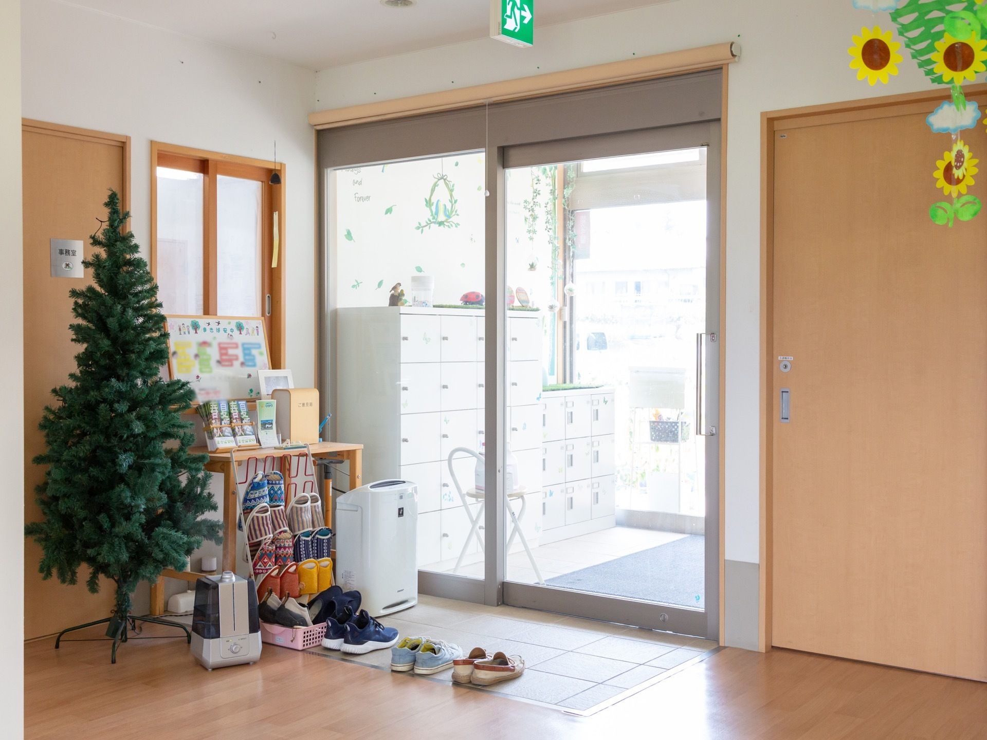 玄関を入るとすぐに事務室がある。玄関の扉は引き戸になっていて近くには空気清浄機、スリッパ立て、加湿器、もみの木などが置かれている。