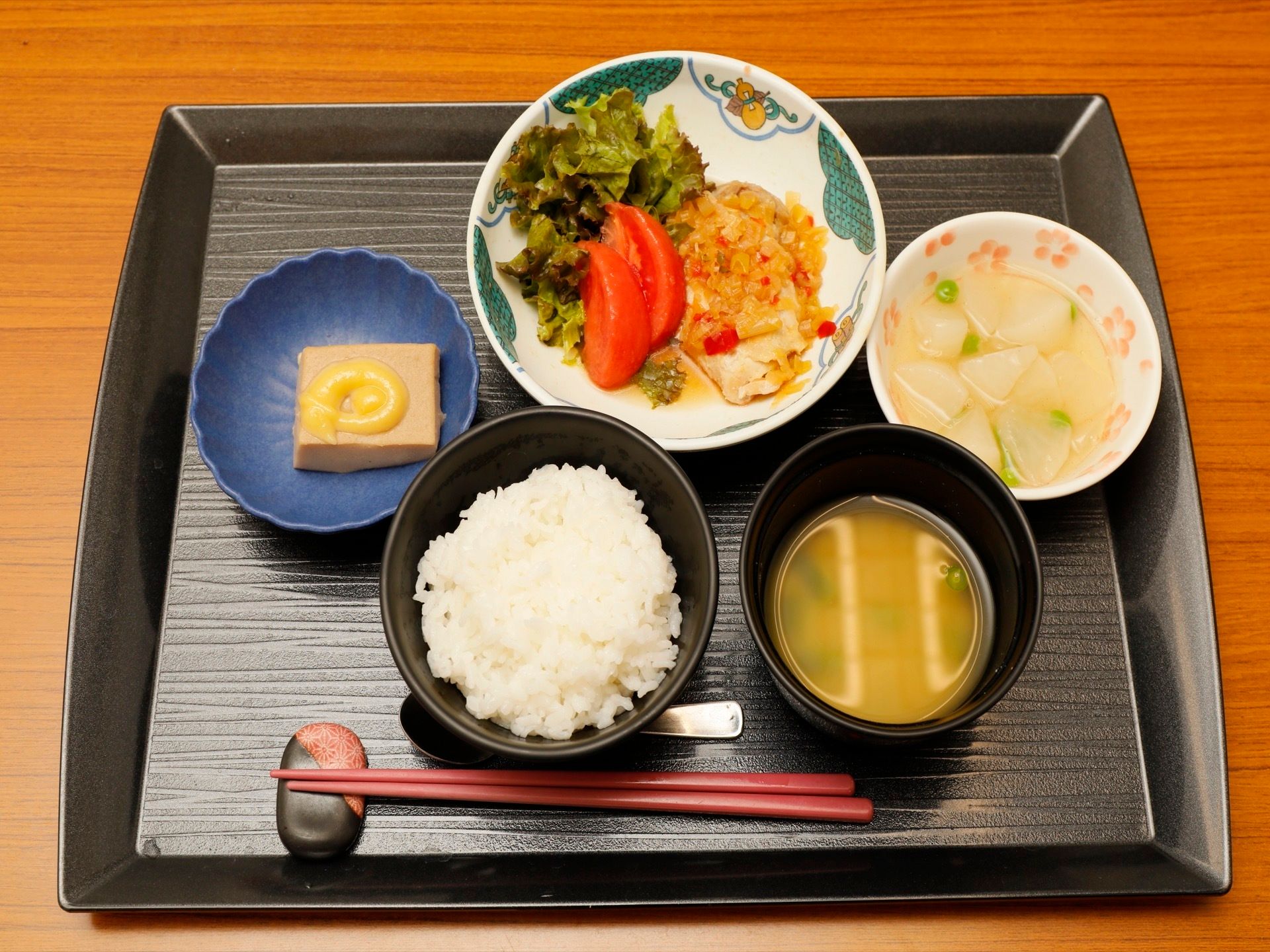 お盆の上にごはんや味噌汁、酢味噌のついた豆腐、トマトや大根で作られたおかずと箸、スプーンが乗せられている。