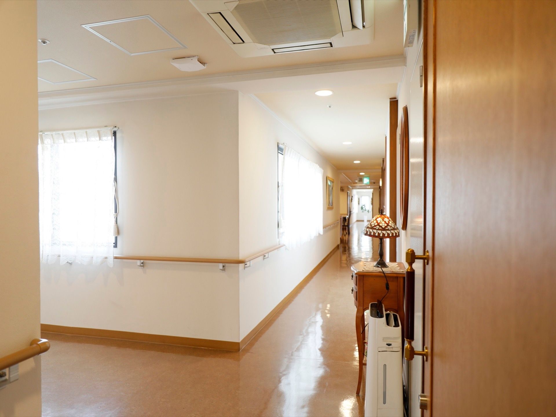 ベージュ色の長い手すりが、白い壁に設置されている。通路の右側には、おしゃれなテーブルランプや空気清浄機がある。