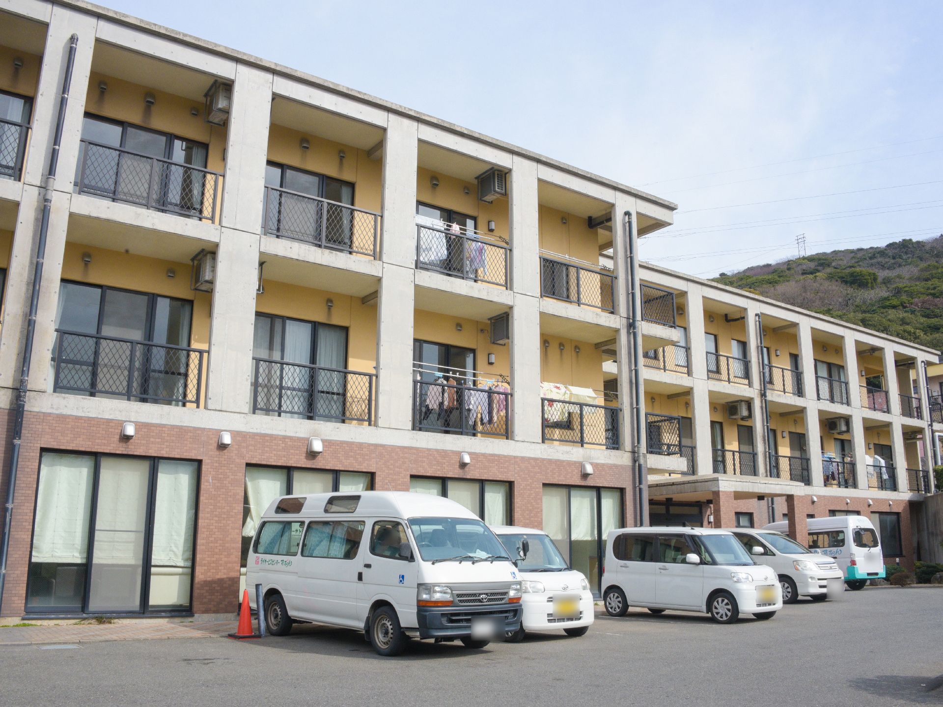 施設は三階建てで、二階と三階部分の外壁は黄色である。黒いフェンスがベランダについている。駐車スペースがあり、車が数台駐車している。