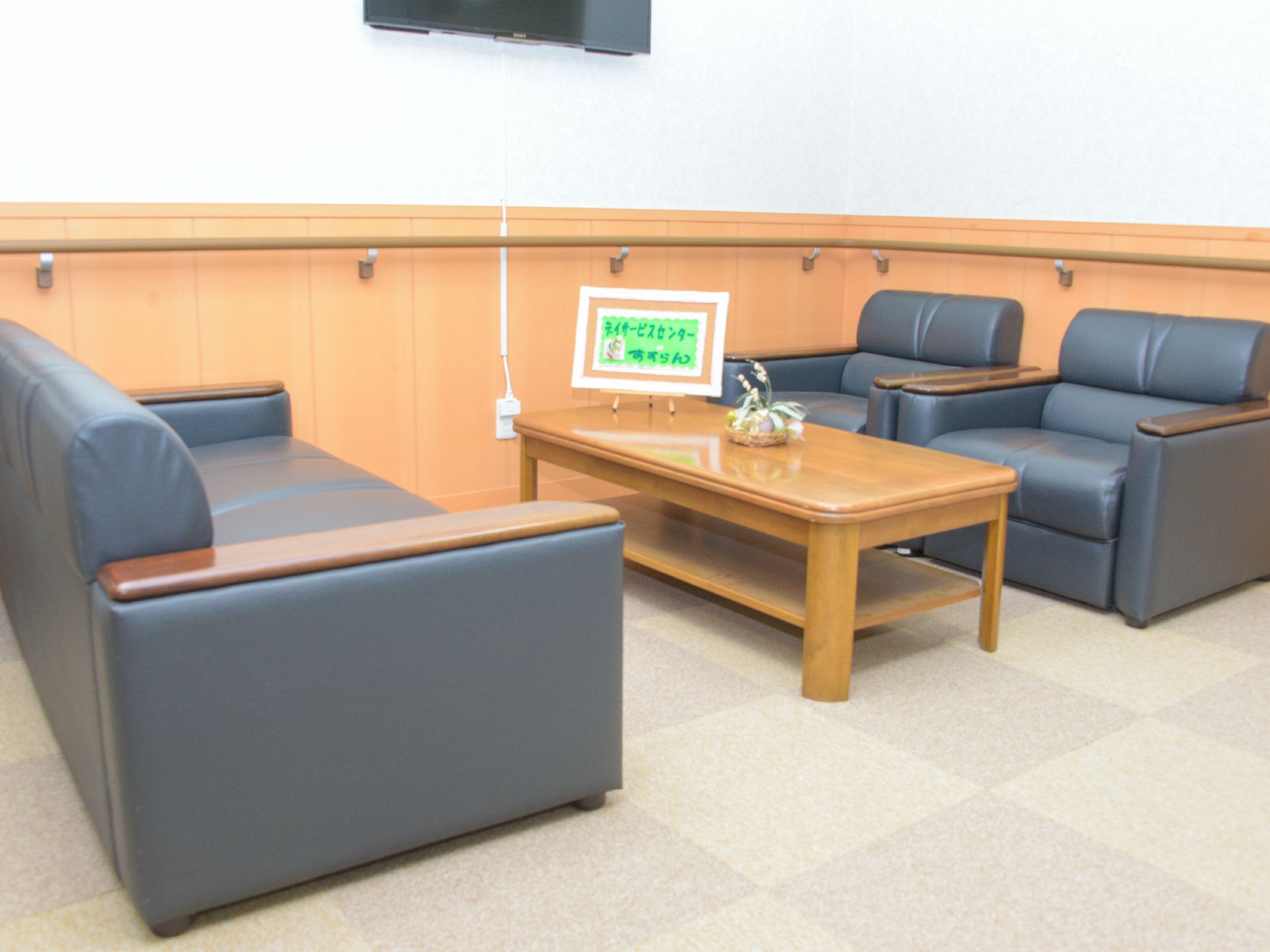 青いアームチェアが置いてある。壁にはテレビが取り付けてある。木製のテーブルの植に、施設の名前がついた額がある。