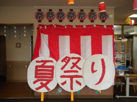 紅白の幕が掛けられ賑やかな雰囲気がある。祭提灯や、大きなうちわに夏祭りと書かれたものが貼られている。