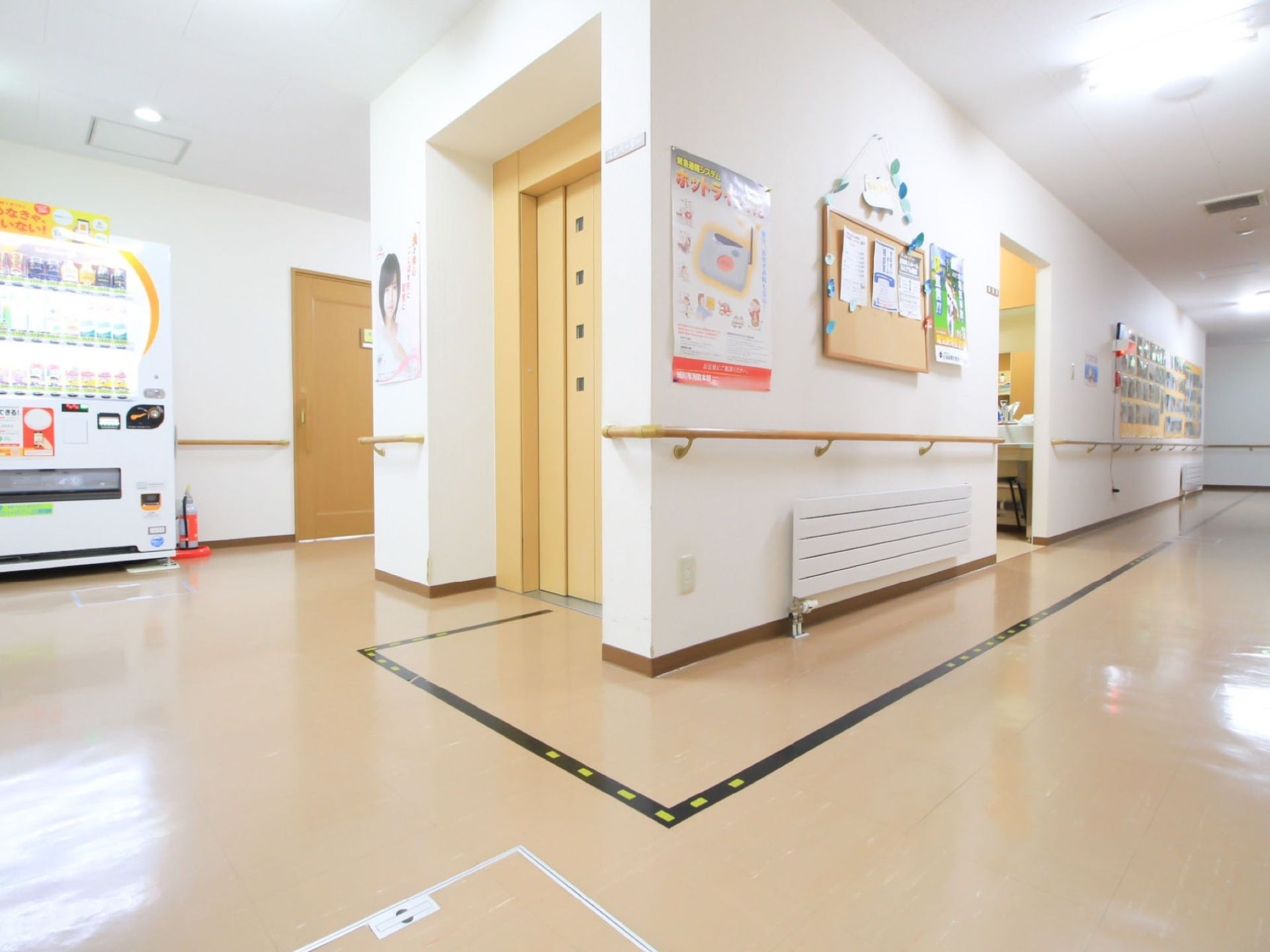 右奥に廊下が伸びている。中央にエレベーターがあり、その裏へ向かって続いている。左奥に自動販売機がある。