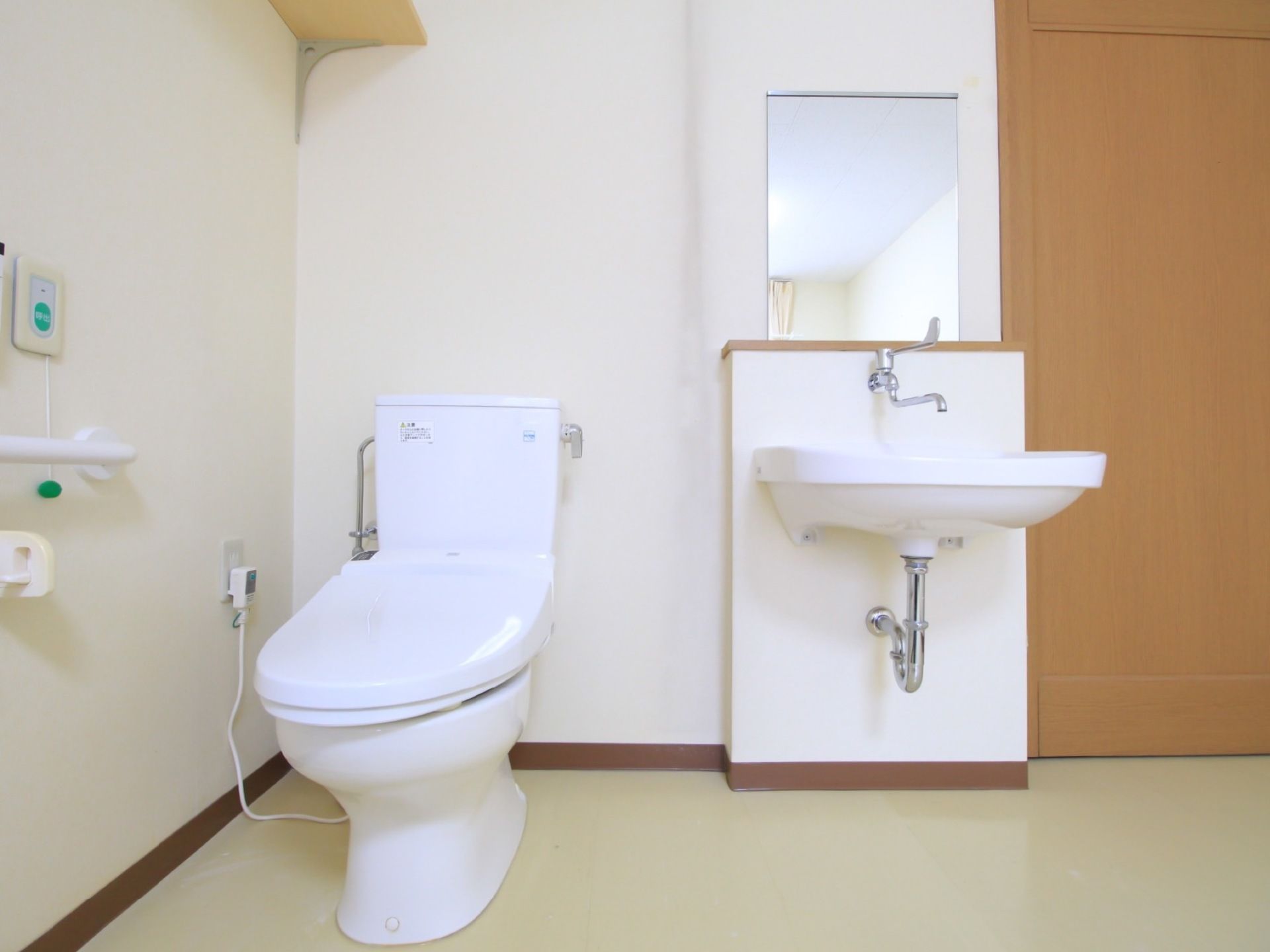 左の壁際に洋式トイレがある。中央に洗面台があり、壁には鏡が取り付けられている。非常ボタンと手すりが用意されている。
