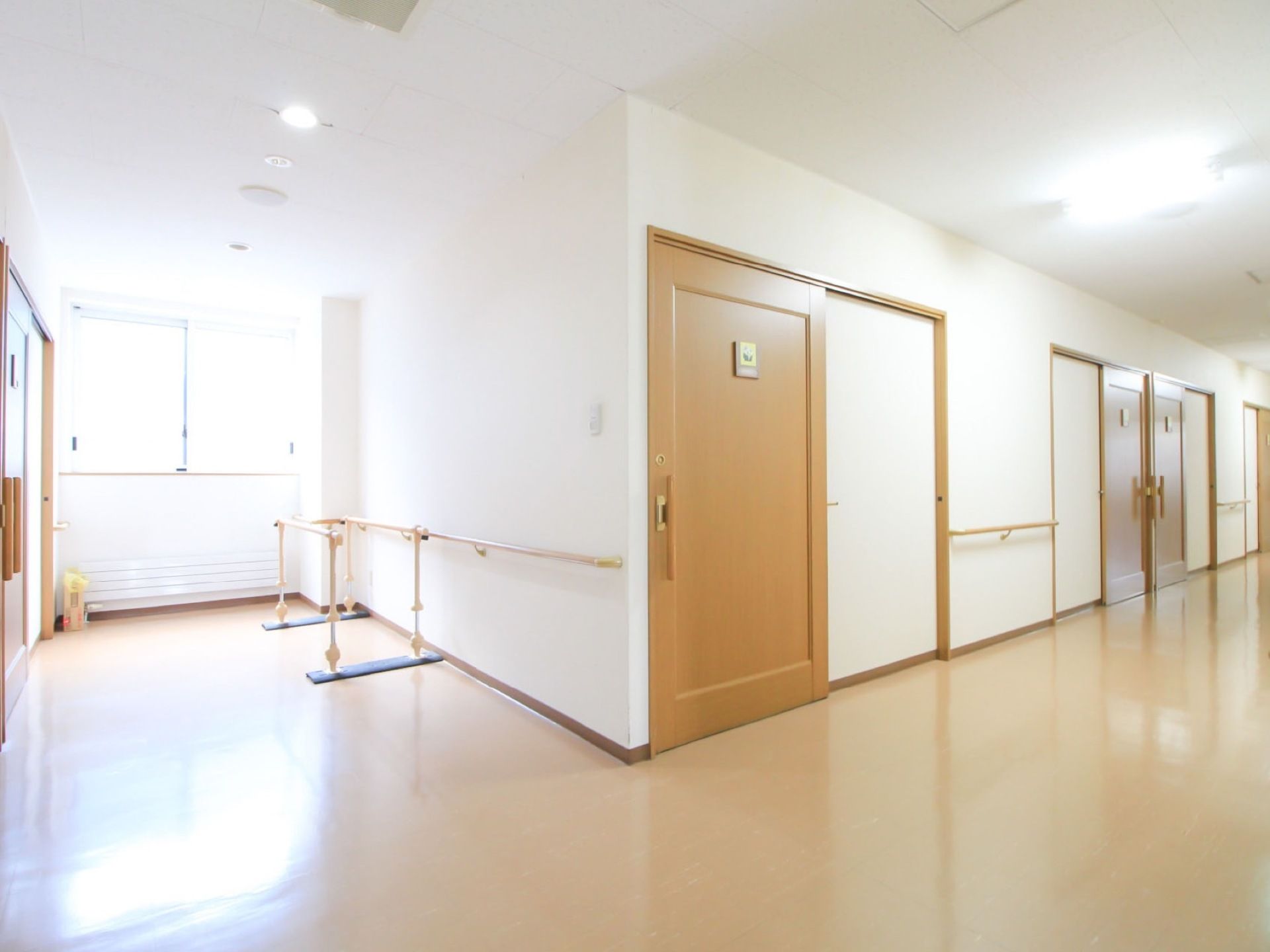 L字型に曲がった廊下で、居室へ続く引き戸が並んでいる。左奥はすぐ突き当りになっており、歩行訓練機が置かれている。