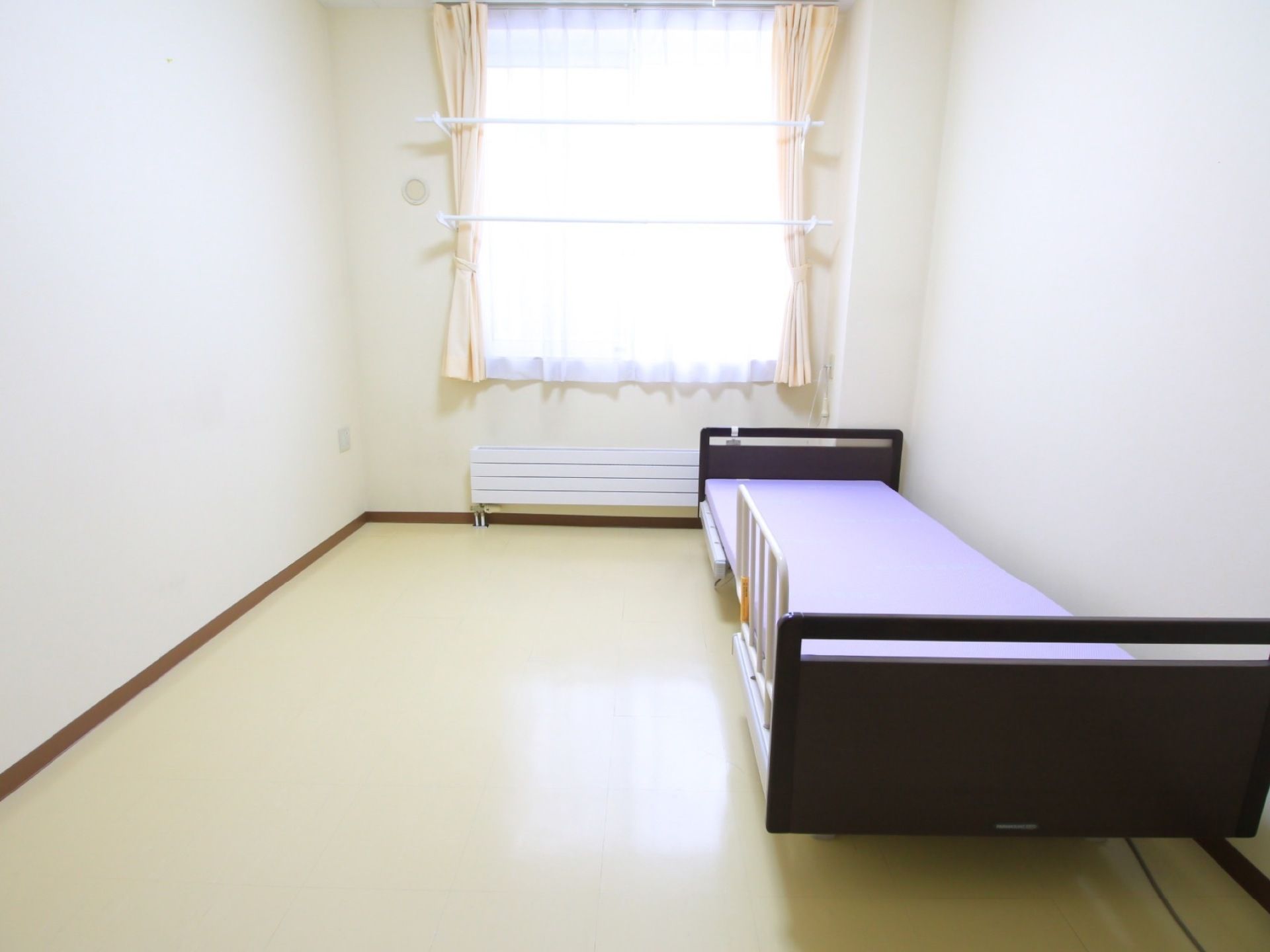 薄いベージュのタイル床の居室で、長方形の間取りである。中央奥にカーテンが閉められた窓があり、レースカーテンが閉められている。