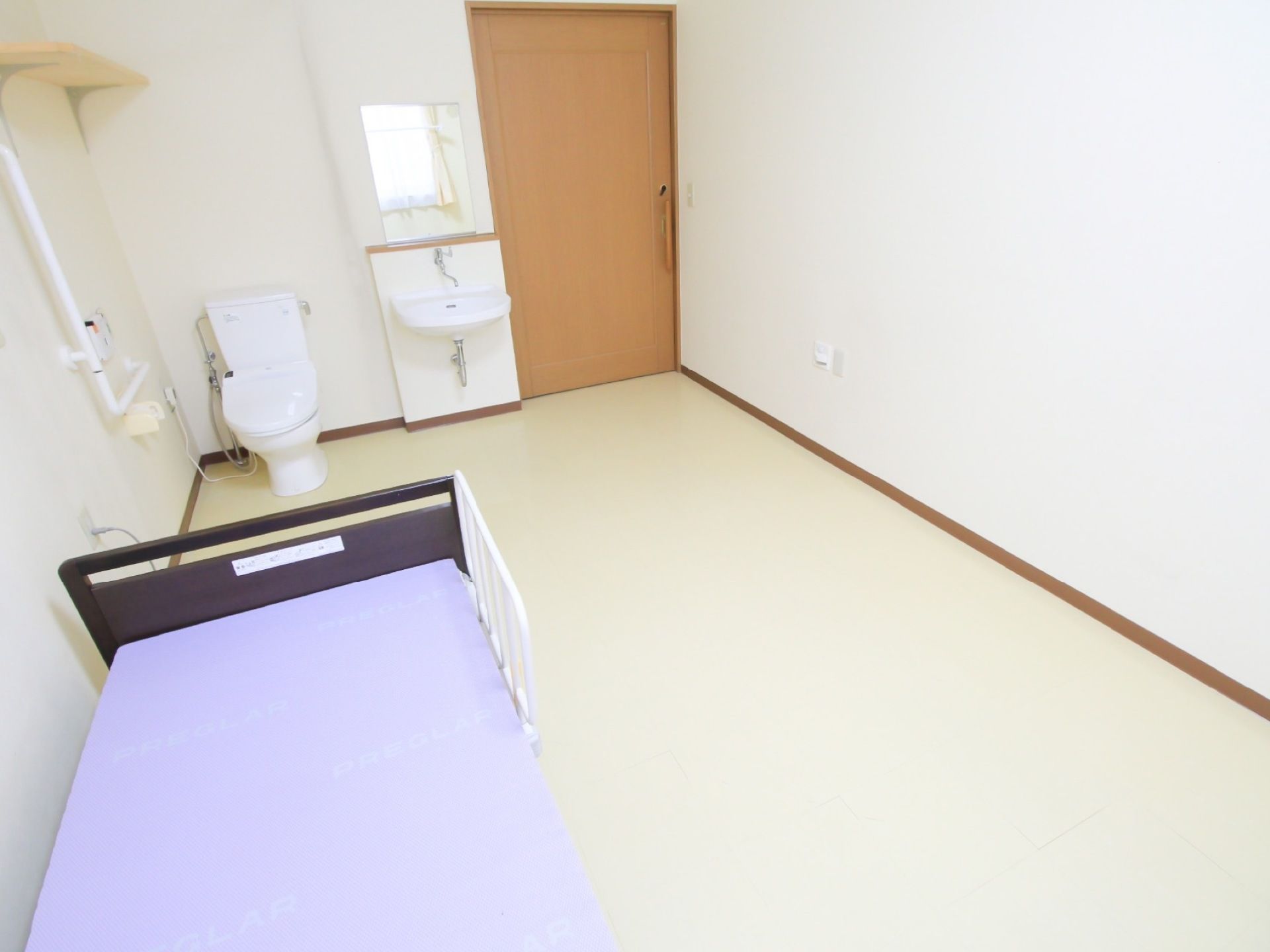 左手前に介護用ベッドがあり、紫色のマットが敷かれている。奥の壁際に洋式トイレと洗面台がある。