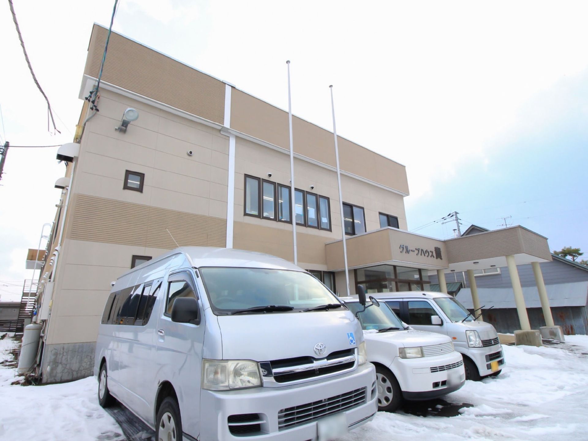 茶色の２階建ての建物が奥にあり、その前は駐車場になっている。施設所有のワゴンなど、車が３台停まっている。