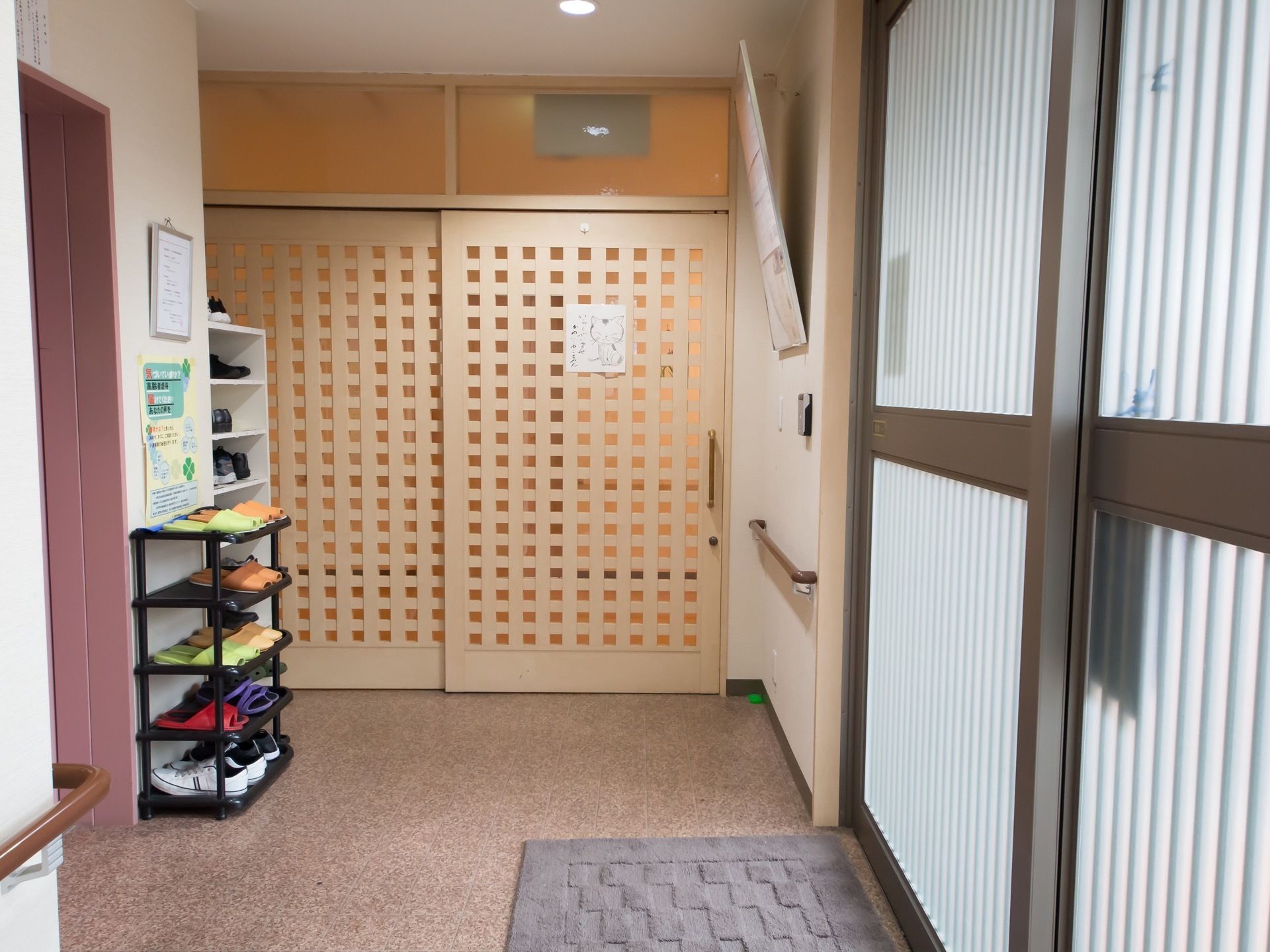 壁には手すりが設置されている。靴を入れる白い棚とスリッパラックが置かれている。奥には猫の絵が描かれた紙が貼ってあるドアがある。