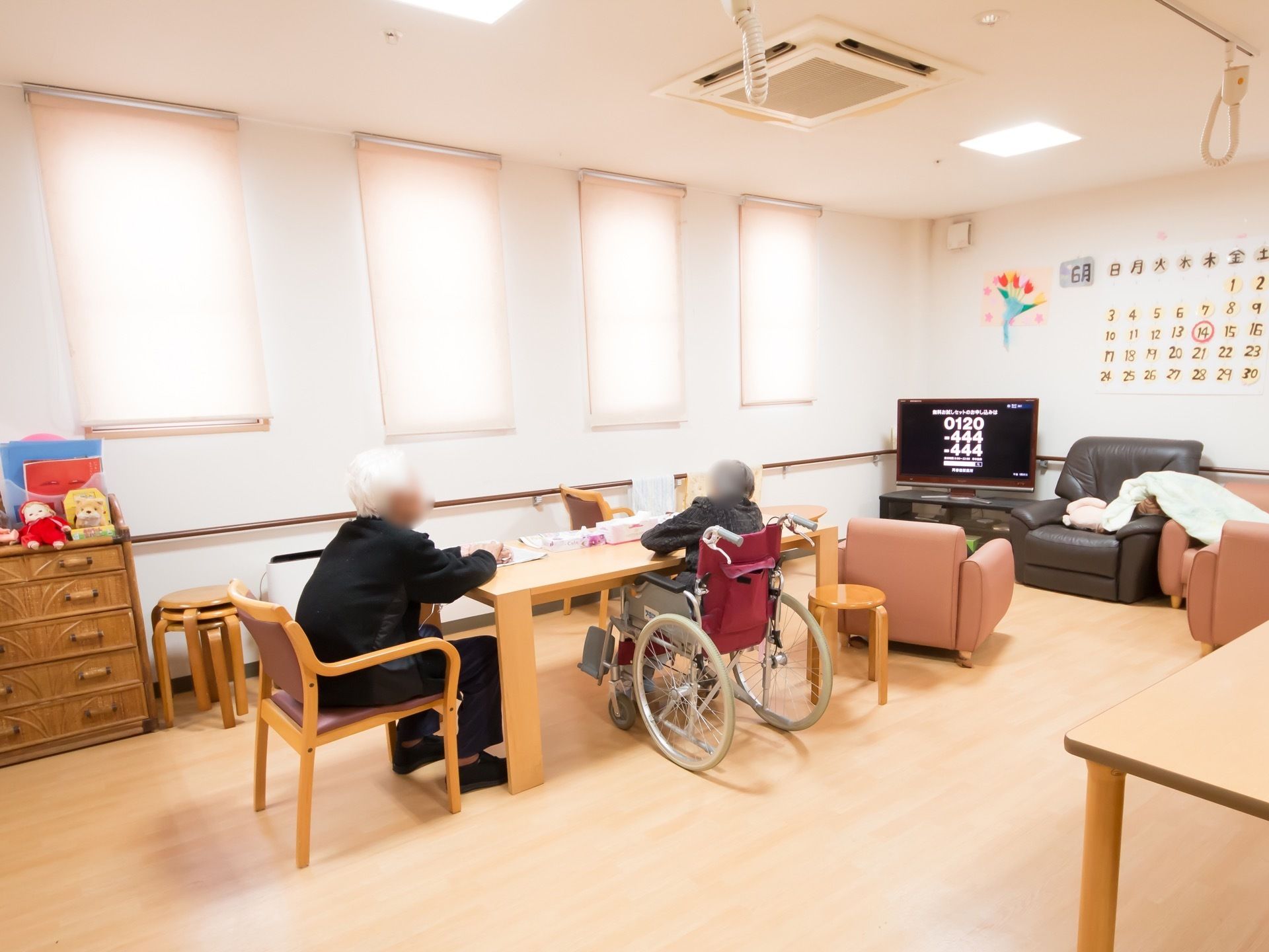 テーブルの前に2人の入居者様が座られており、テレビをご覧になられている。壁には手すりが設置されている。