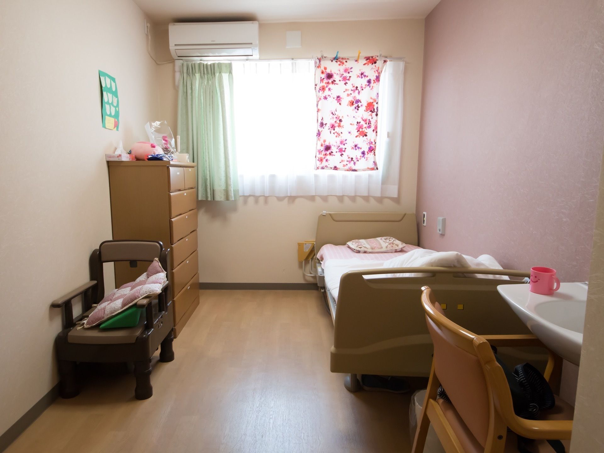 壁側にベッドが1台置かれている。窓には緑色のカーテンが掛けられており、上部にはエアコンが設置されている。