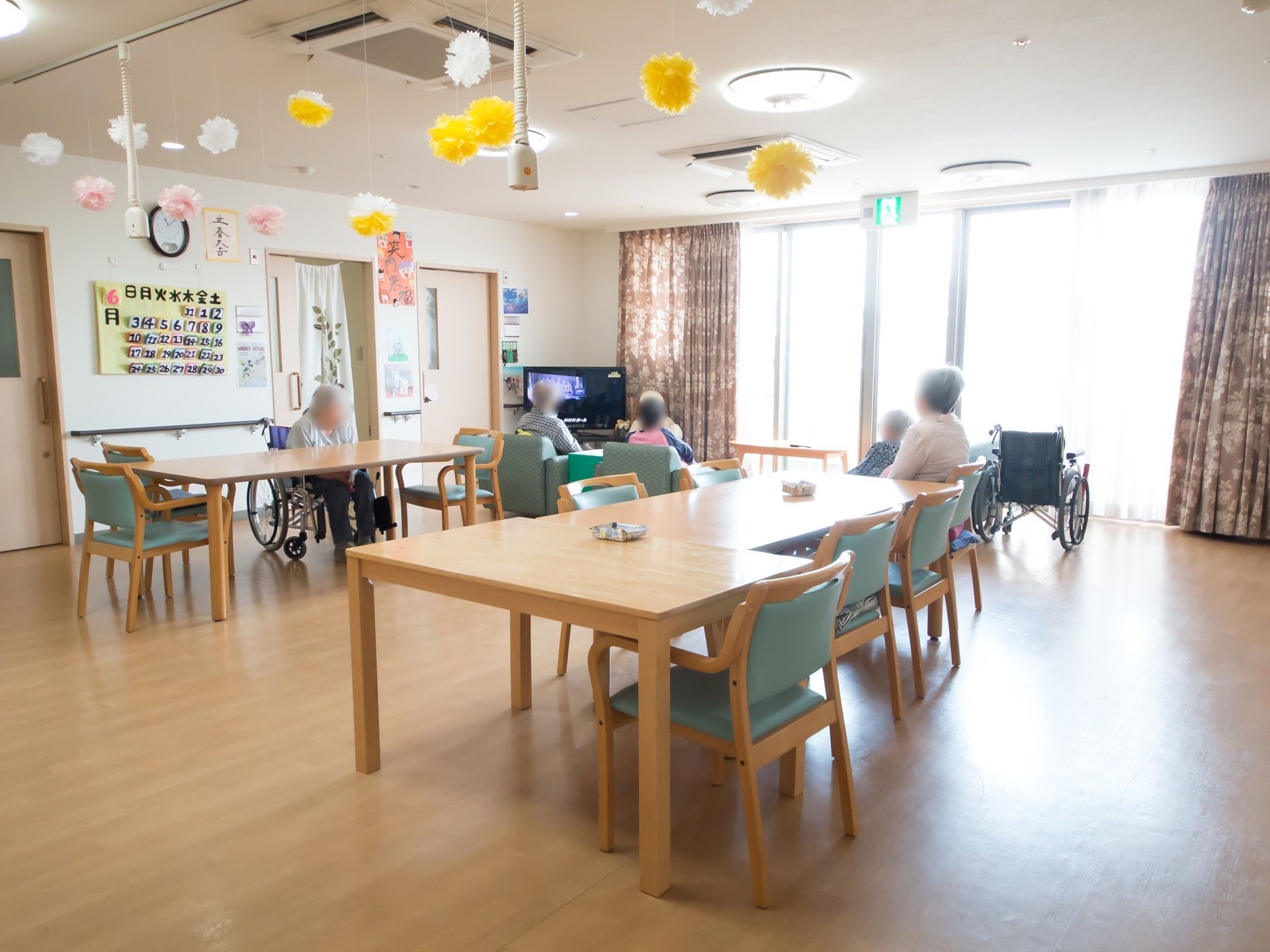 壁には手作りのカレンダーが飾られている。テレビの前に複数の入居者様が集まりテレビをご覧になられている。