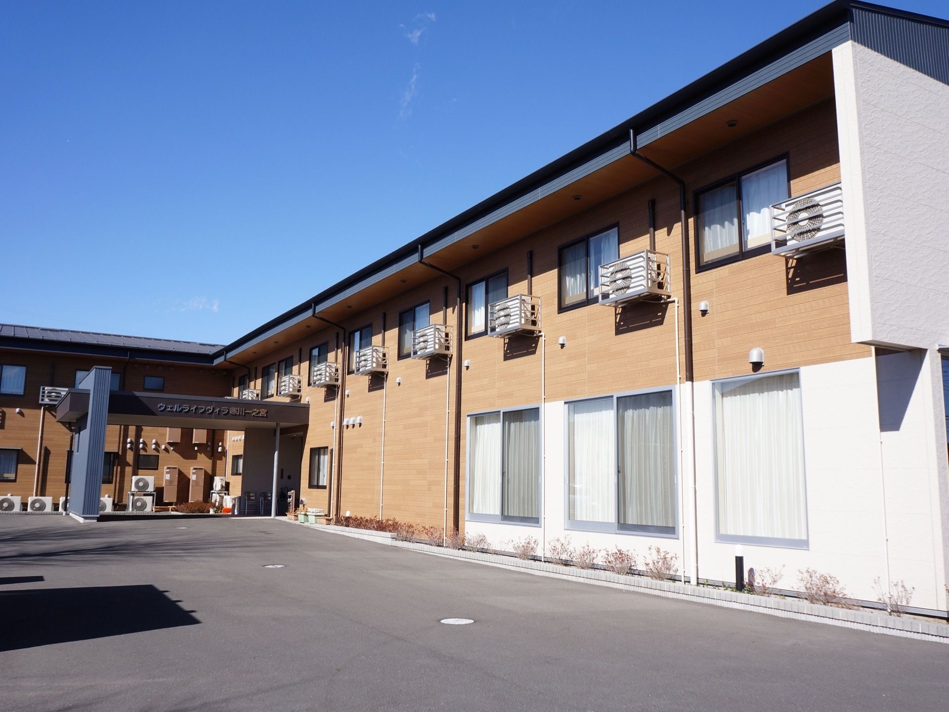 白と木目調を基調とした２階建ての建物がＬ字に建っている。建物には、たくさんの室外機が設置されている。