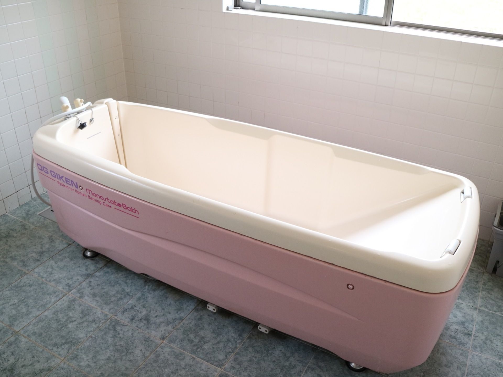 窓が付いた広いタイル張りの浴室である。介護用特殊入浴装置の一部であるピンク色の浴槽が設置されている。