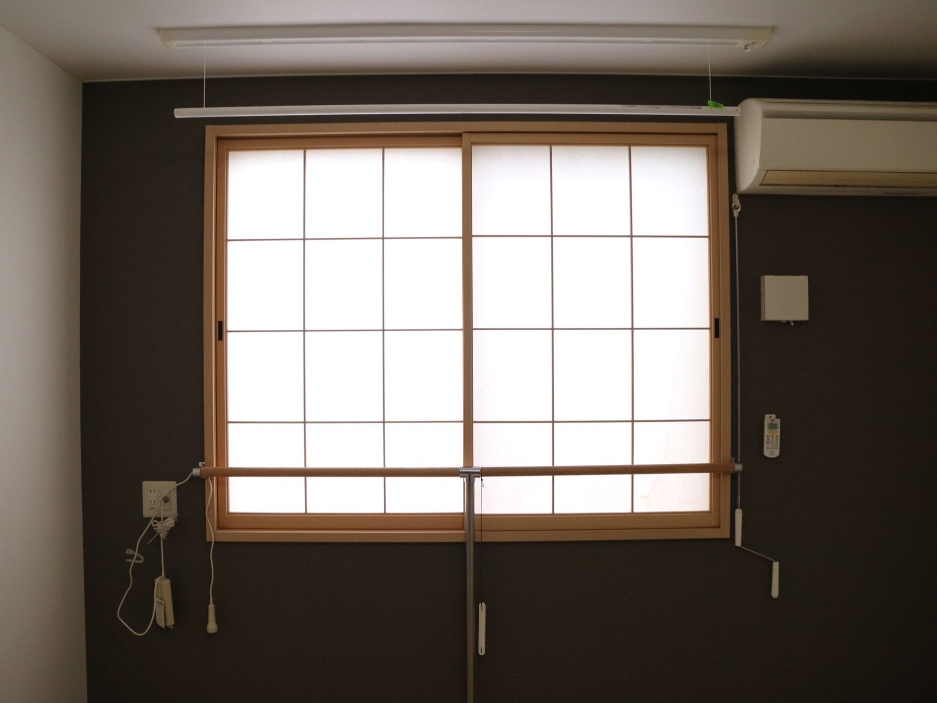焦げ茶色の壁面に窓とエアコンが設置されている。窓には、障子が取り付けてあり、その手前にＴ字型の棒が取り付けてある。