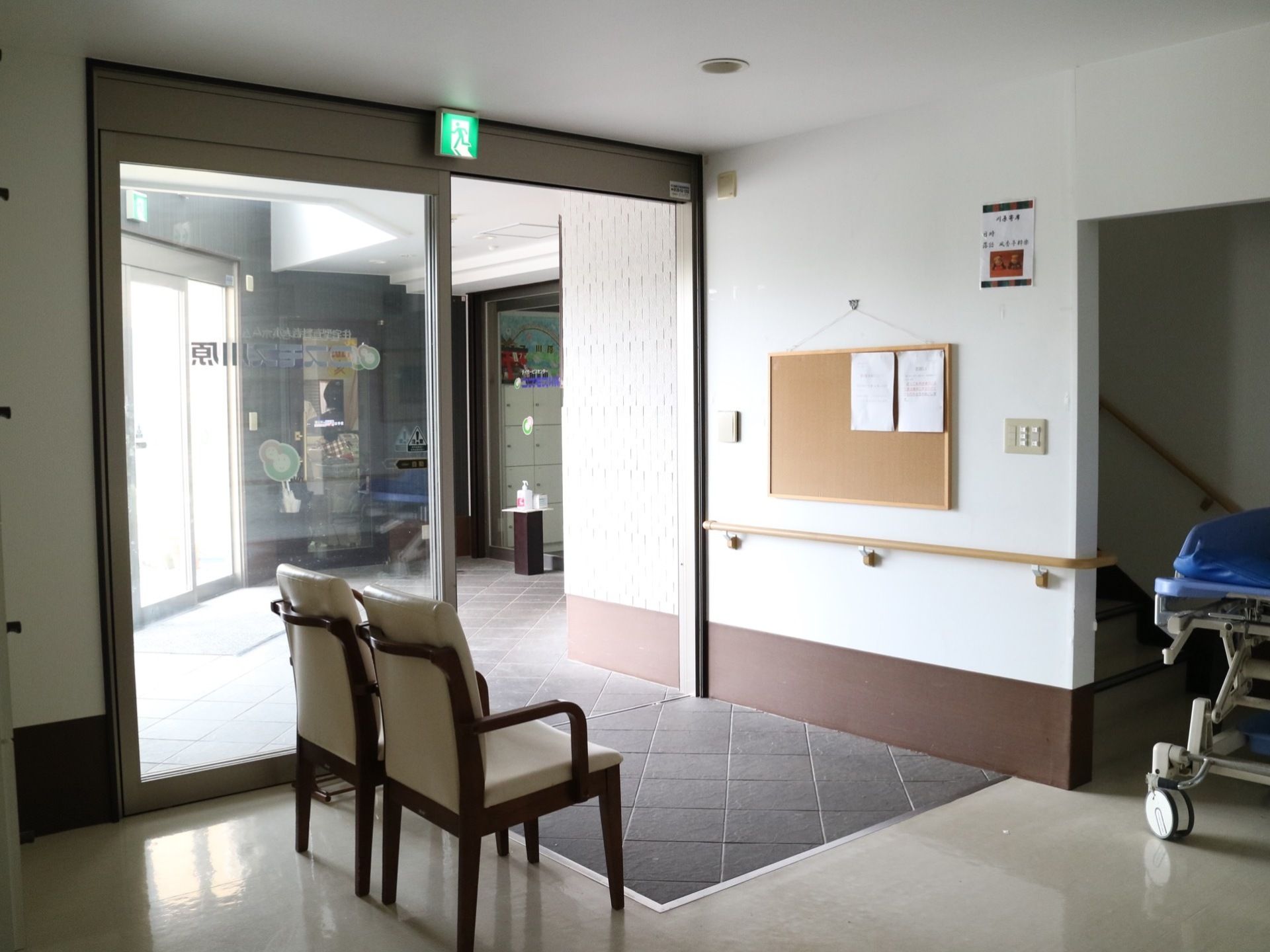 正面玄関から斜めに位置する場所に自動ドアが設置されており、背もたれの高い椅子が２脚置いてある。階段やストレッチャーが見える。