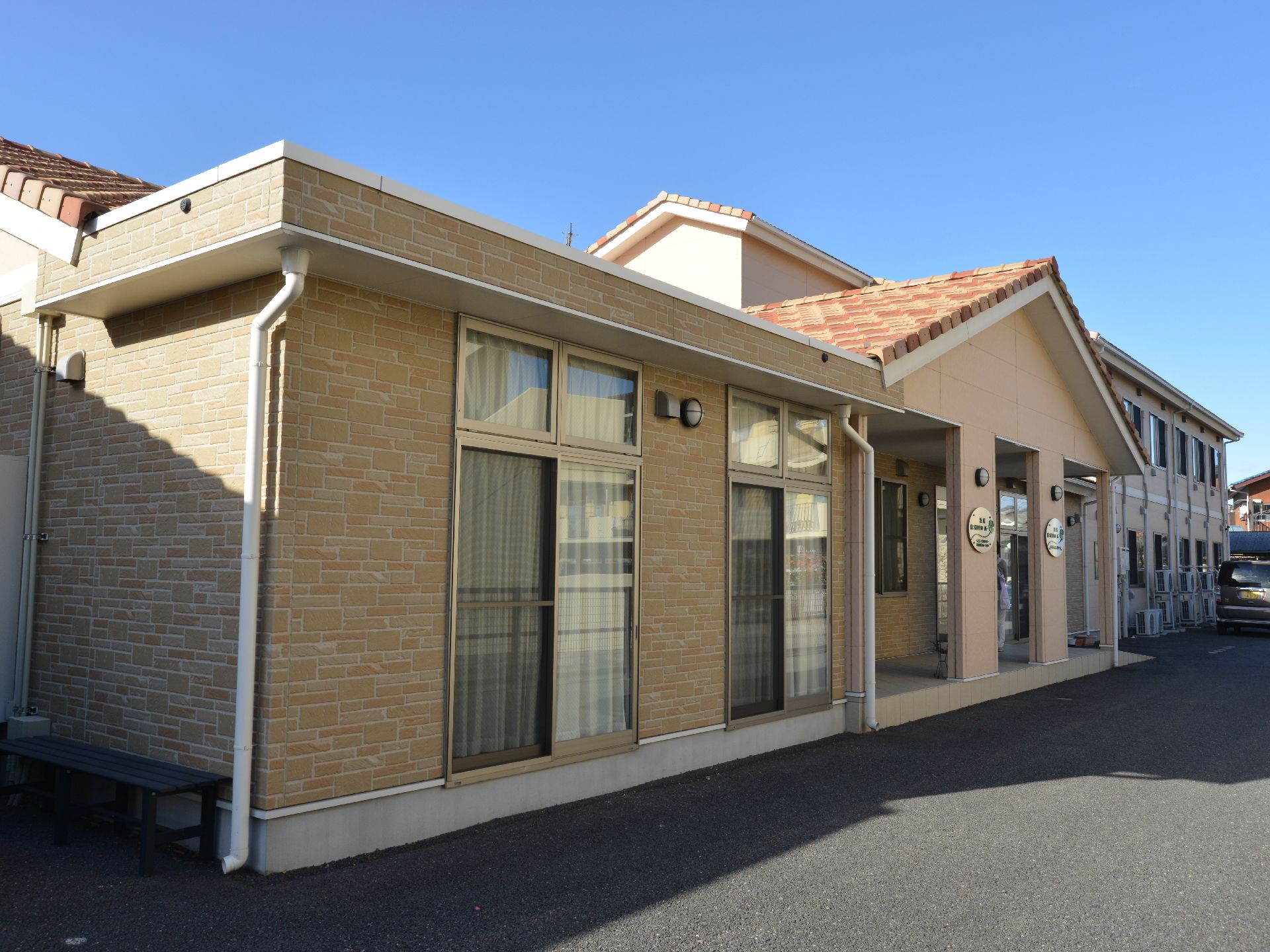 ベージュが基調となっている建物は、暖色系の瓦屋根の和風なデザインになっている。建物の外には駐車場が。
