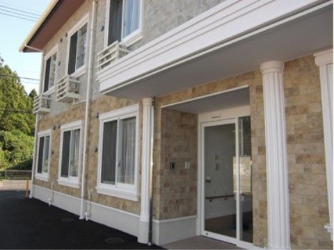 レンガ調の外壁が特徴的な施設建物である。右側には屋根付きの玄関がある。左側には木が立っており、自然が近くにある環境である。