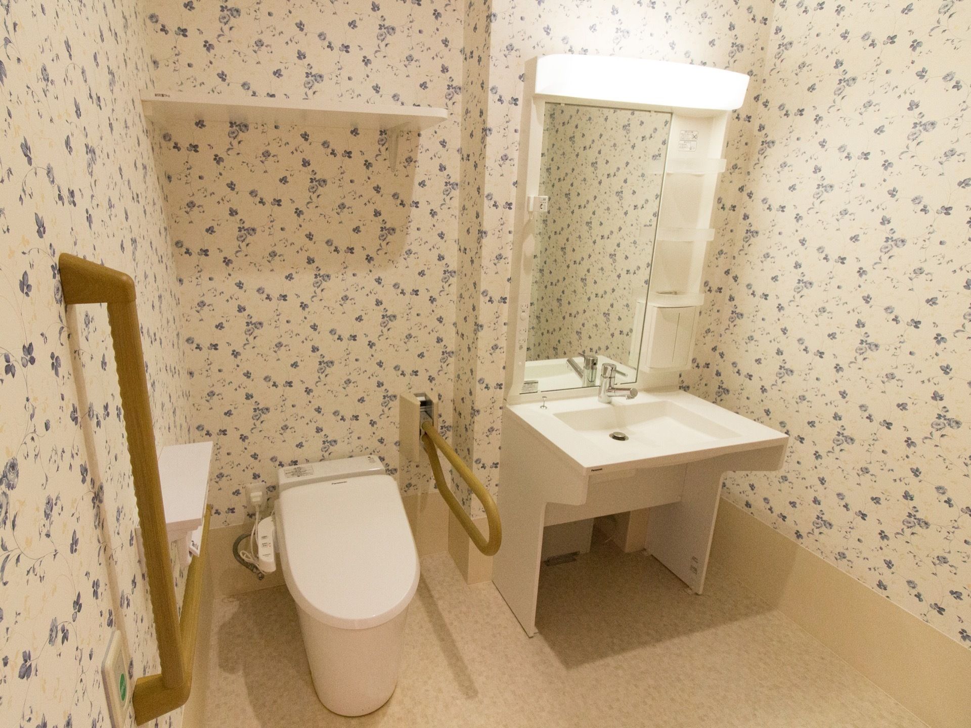 花柄の壁紙が明るくおしゃれなトイレになっている。手すりと洗面台が設置されており、車椅子でも楽に移動できる広さがある。