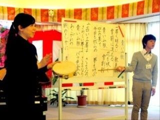 スタッフが2人立っている。ホワイトボードには紙が貼られており、そこを棒で指している。スタッフの1人はタンバリンを持ち、リズムをとっている。