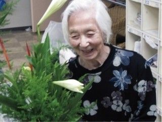 入居者様が花を見て微笑んでいる。後ろには白い棚が置かれており、床は格子柄になったカーペットが敷かれている。