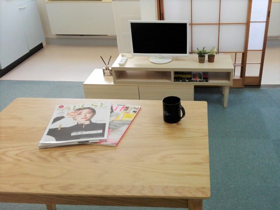 雑誌が２冊と黒いマグカップが置かれた木製のテーブル。真正面にはテレビが置かれたテレビラックが置かれている。