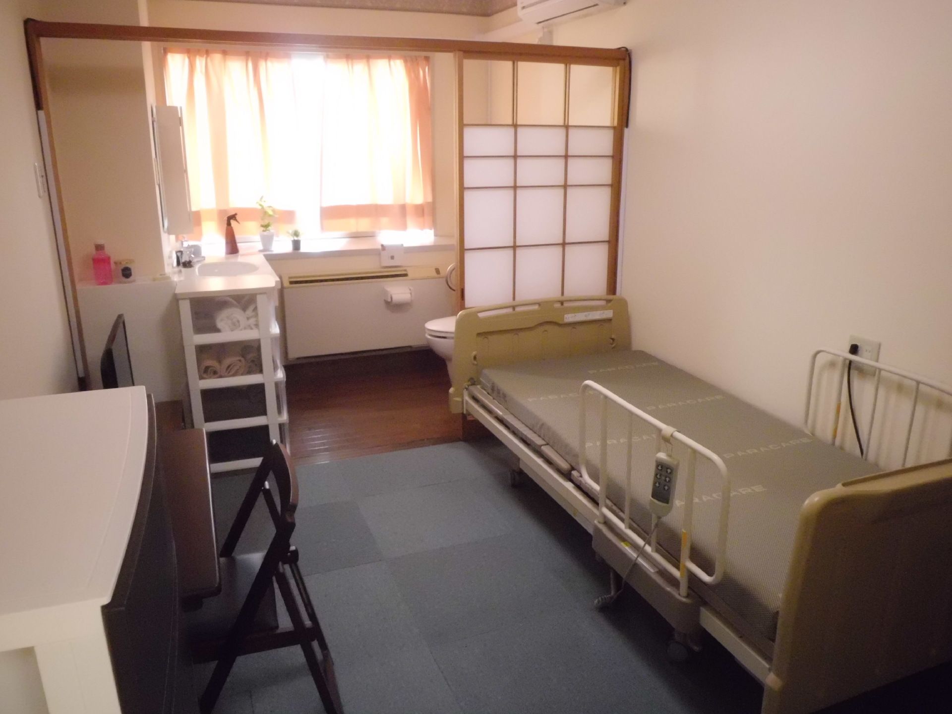 介護ベッドが右手、テレビやテーブルセットが左手、そして奥のフローリングのスペースにはトイレがある。