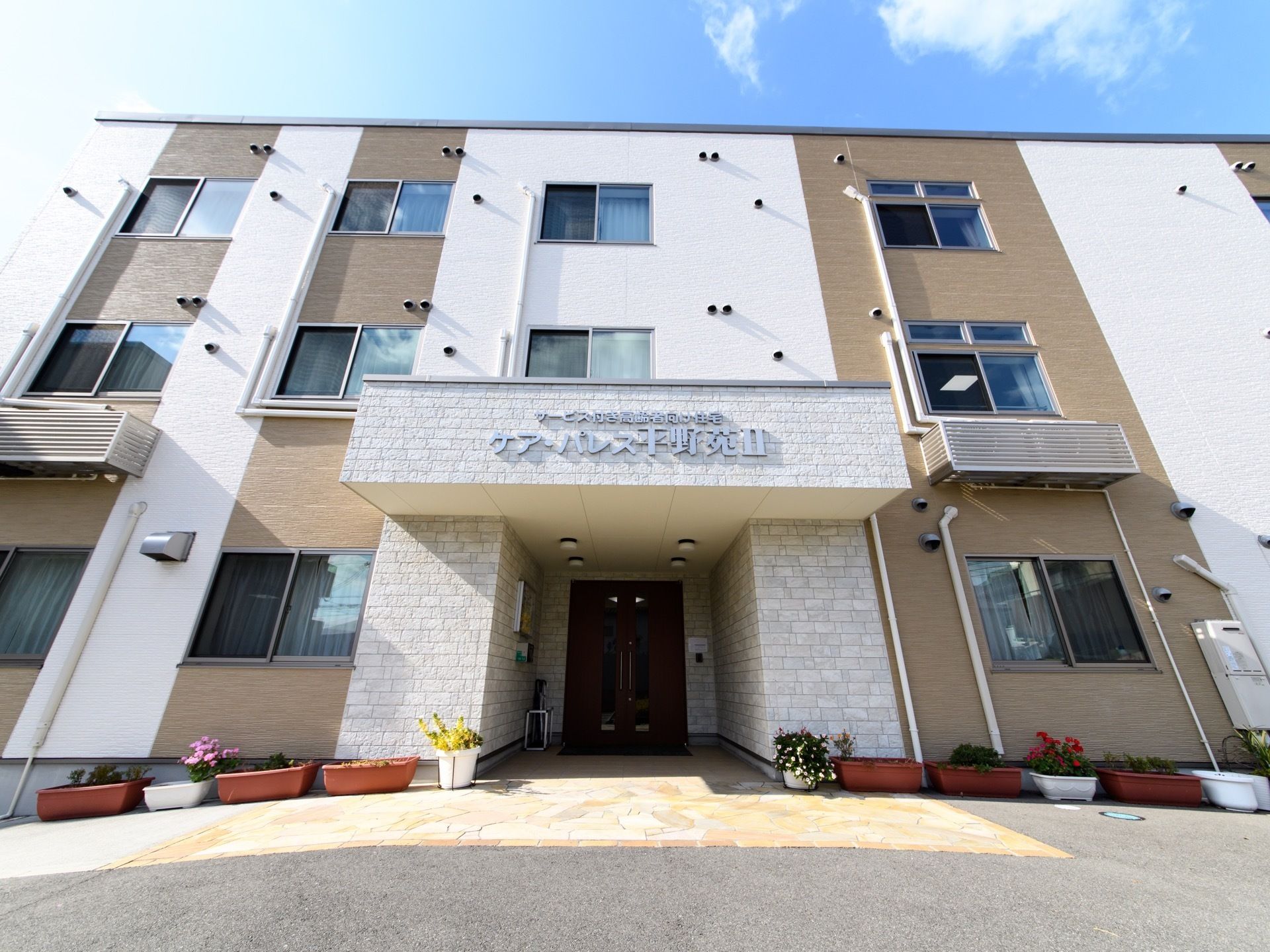 施設は3階建ての建物。建物前にはさまざまな花が植えられたプランターが複数ある。入口前はひさしがあり、傘立てが置かれている。