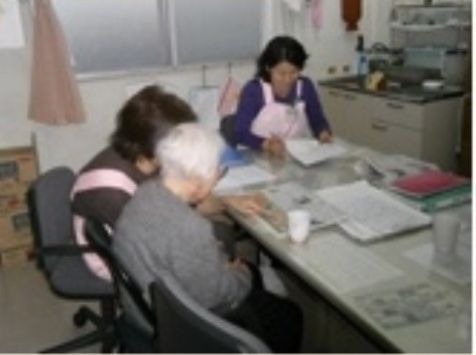 作業テーブルのような上に広げたものを白髪の高齢女性に読んであげている女性