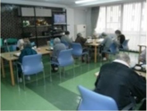 緑の床にテーブルが並んでおり、青い椅子に腰かけて高齢者が食事をしている食堂