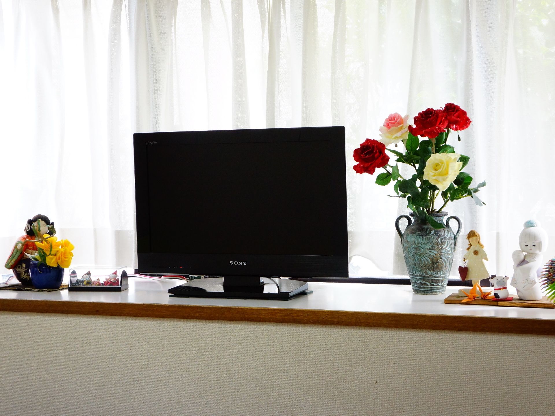 光の差し込む出窓には白いカーテンがかかり、テレビが設置されている。その周辺には花や、人形など色とりどりの置物が飾られている。